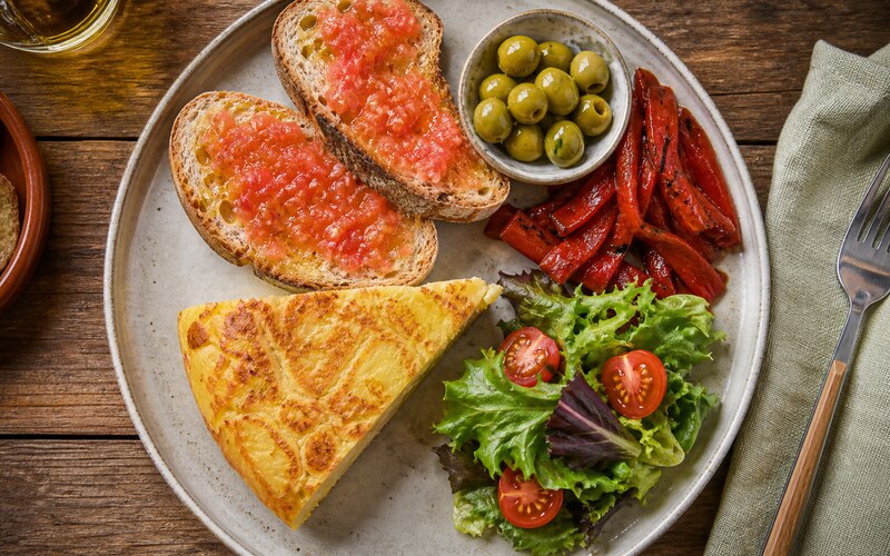 Plato español visto desde arriba con tortilla, pan con tomate, pimientos y aceitunas